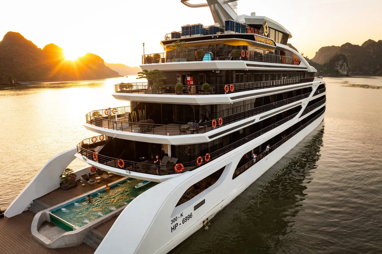 A white multi-deck steel cruise ship with a stern pool and life rings sits on calm water at sunset, with limestone karst islands in the background.
