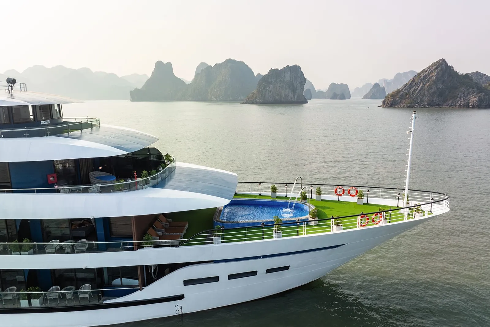A white cruise ship with multiple decks, a swimming pool, and green artificial turf on the sundeck, photographed from aboard the vessel with limestone karst islands visible in the calm bay water.