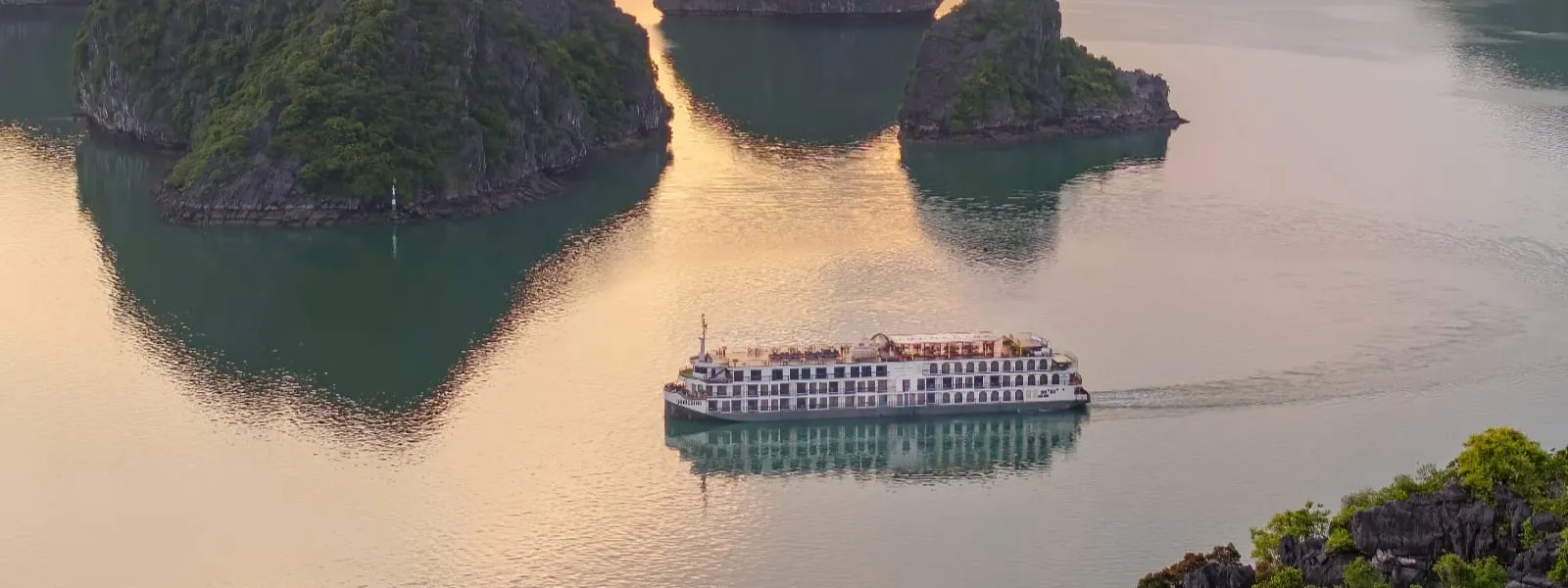 Aerial view of a white multi-deck cruise ship sailing between limestone karst islands on calm, golden-lit water.