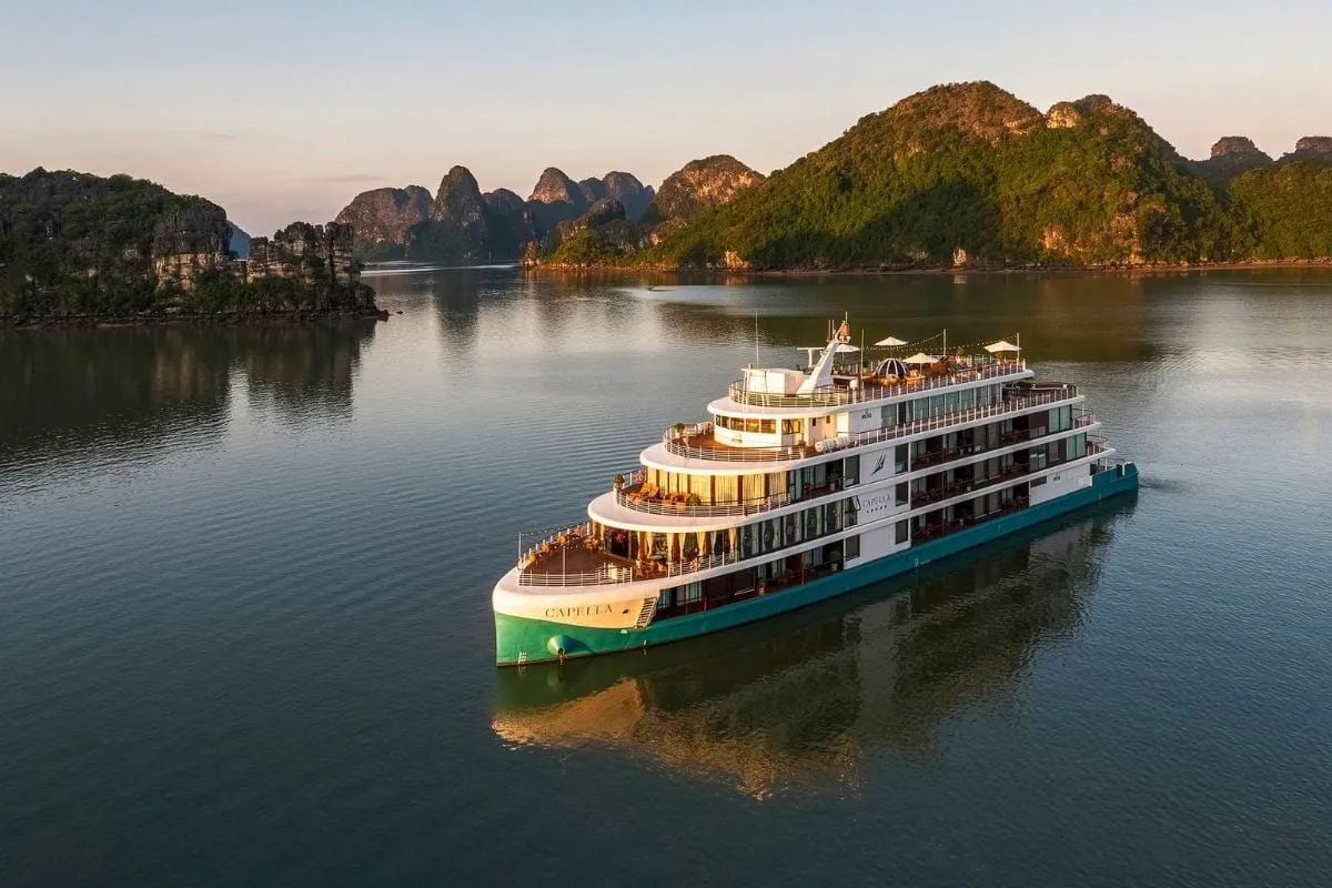 A multi-deck cruise ship with illuminated decks floats in calm water surrounded by dramatic karst limestone mountains at dusk.