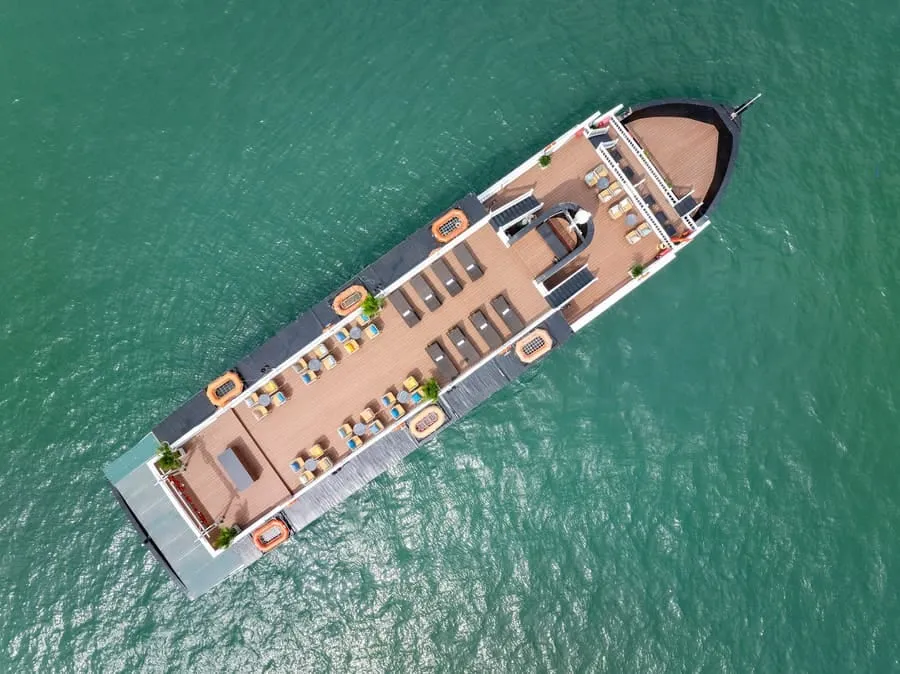 Top-down aerial view of a cruise boat on turquoise water, showing the wooden sundeck with rows of loungers.