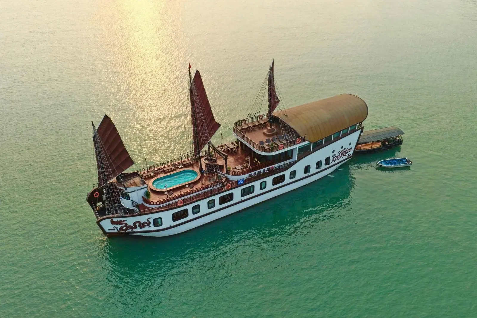 An aerial view of a white junk-style cruise boat with dark red sails, a rooftop pool and a curved golden roof, anchored on calm green water beside a small tender and a speedboat, with a script logo and a dragon painted on the hull.