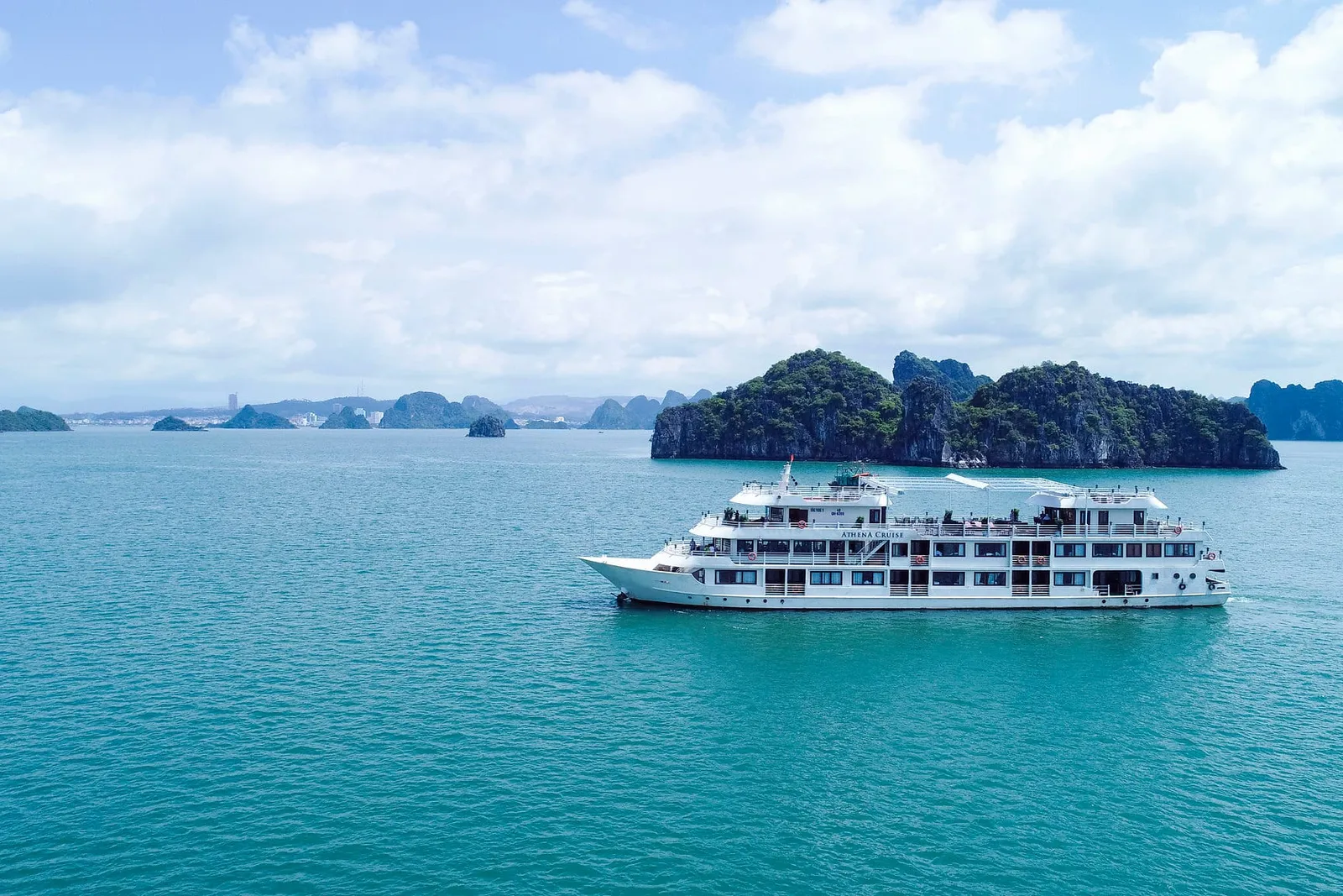 A white multi-deck cruise ship floats in turquoise water with limestone karst islands in the background under a cloudy sky.