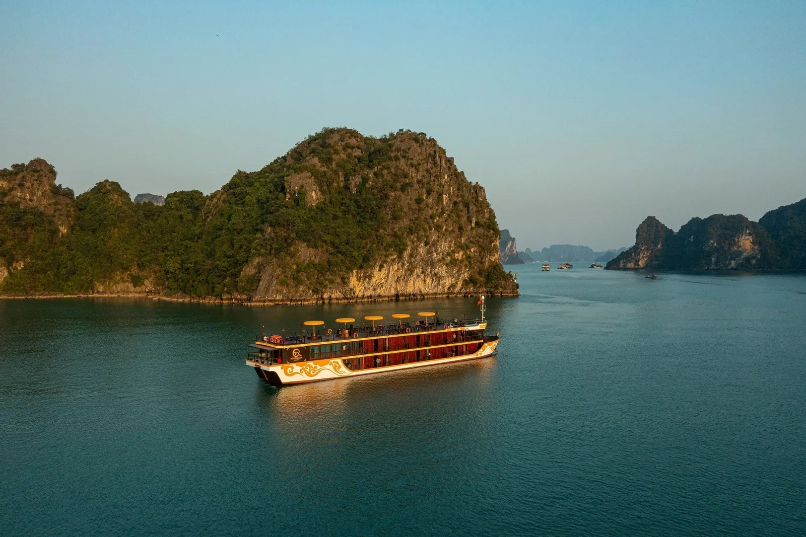 Aerial view of a two-deck wooden-style cruise boat with a cream and gold hull, dark red cabin walls and yellow umbrellas on the top deck, sailing in calm water beside forested limestone karst islands at golden hour.