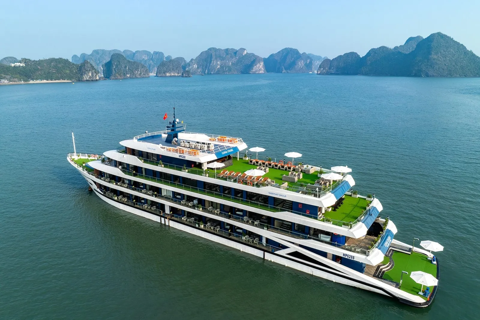 Aerial view of a white and blue multi-deck steel cruise ship with green artificial-turf decks and white parasols, anchored in calm green water with limestone karst islands behind.