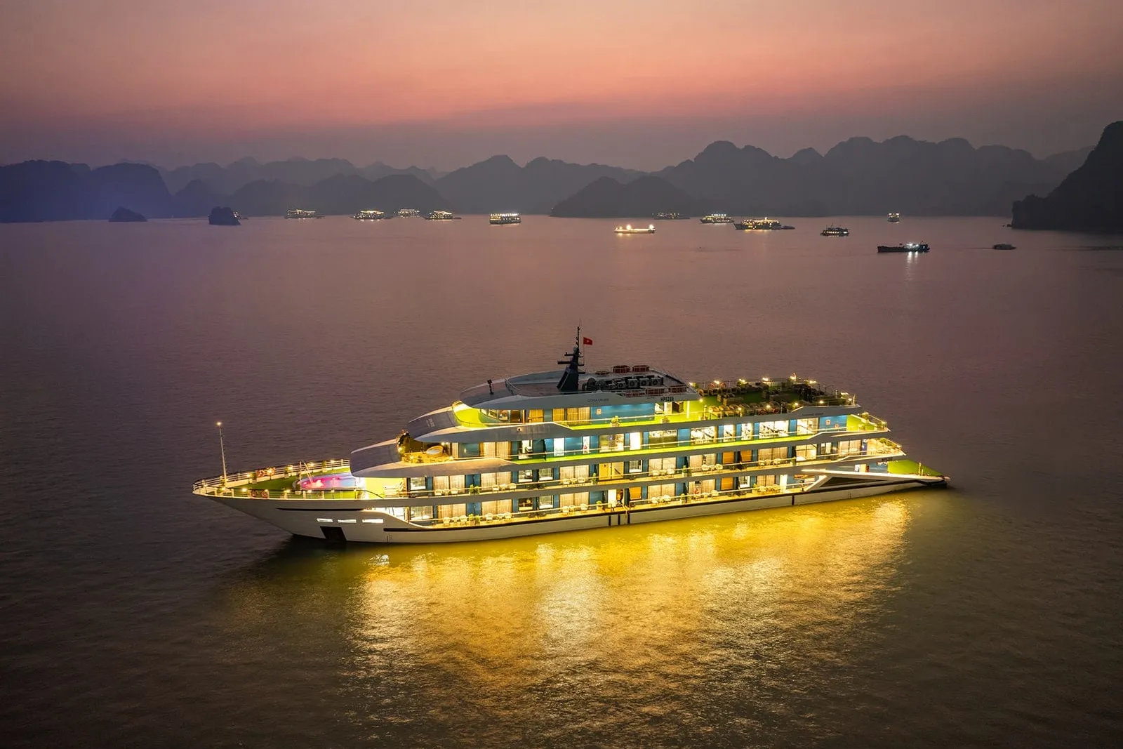 Aerial dusk view of a large white multi-deck cruise ship lit with warm lights, anchored on calm water with limestone karsts and other distant boats under a pink-purple sky.