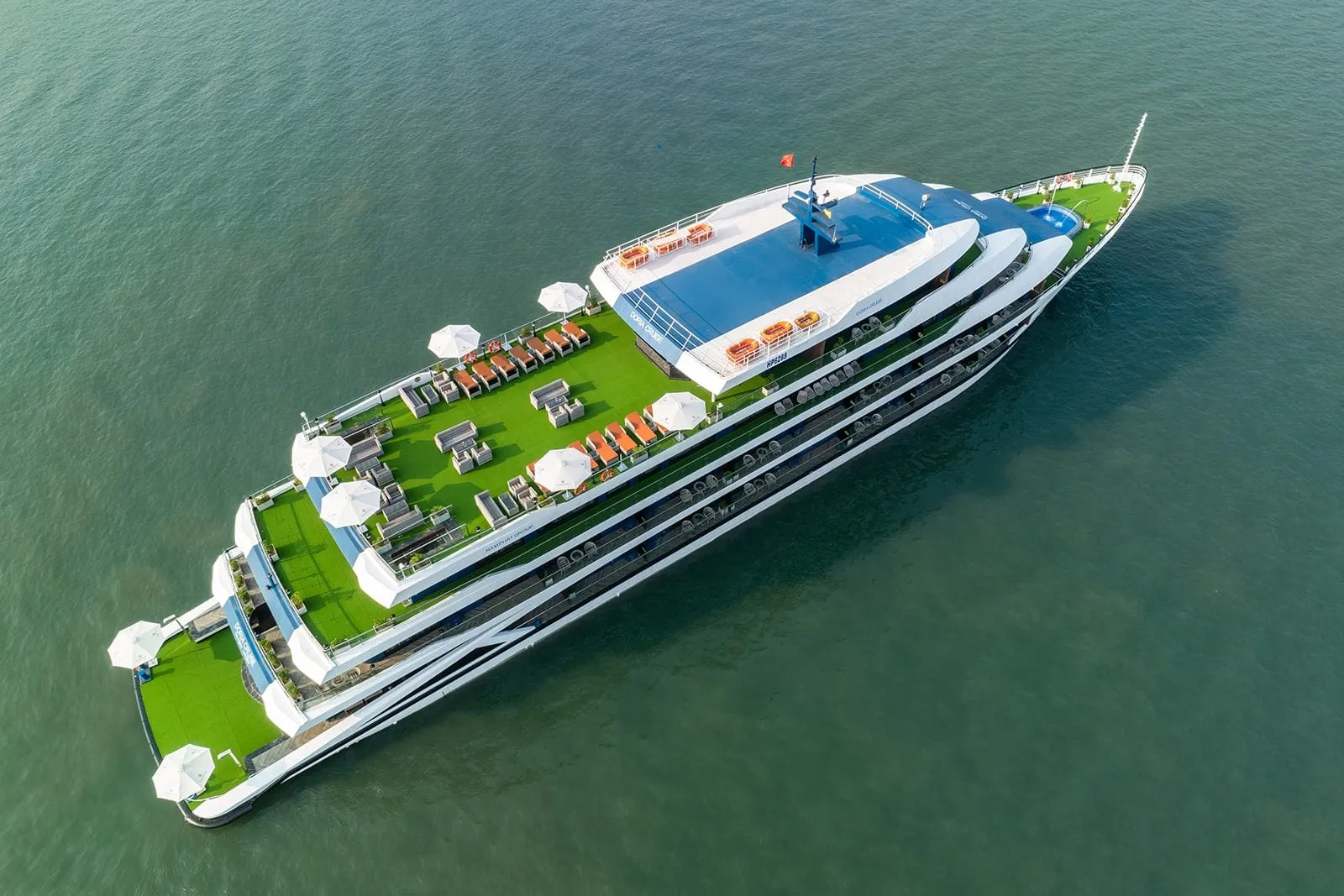 Aerial view of a white steel multi-deck cruise ship with green turf sun decks, white umbrellas, orange loungers and a blue-topped upper deck on calm green water.