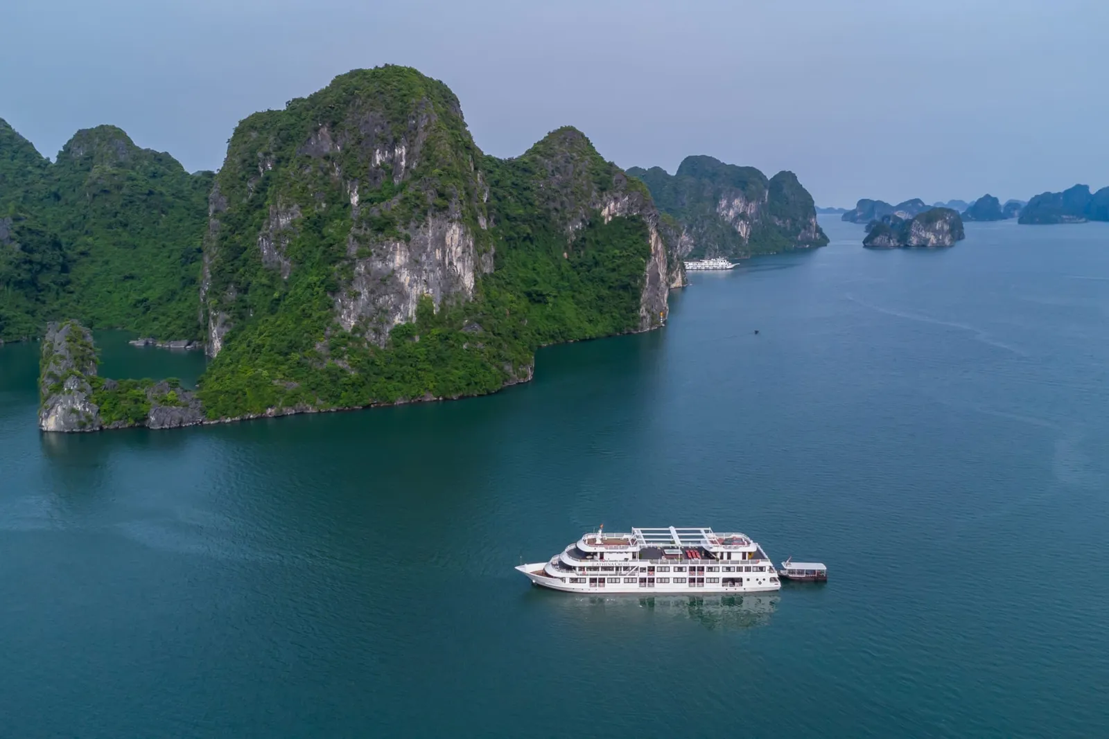 Aerial view of a white multi-deck cruise ship with a small tender boat alongside, anchored in calm green water among forested limestone karst islands, with another white ship in the distance.