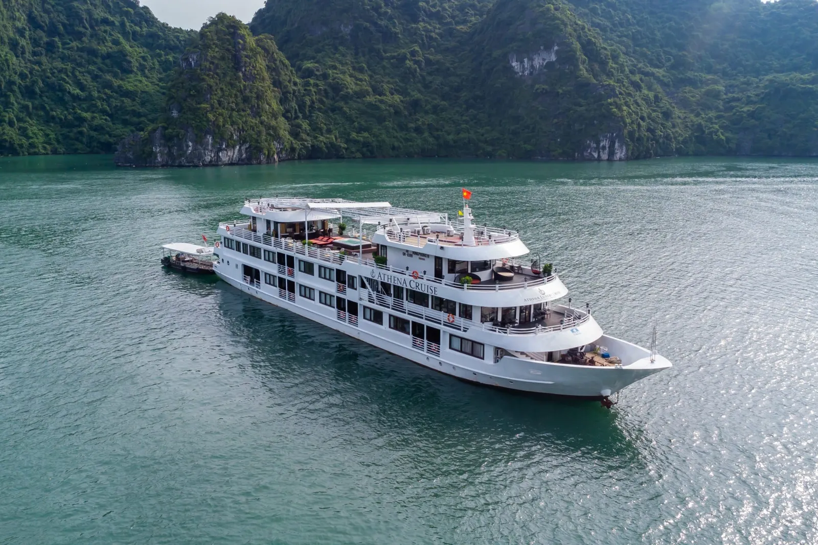 Aerial view of a white multi-deck cruise ship with 'Athena Cruise' on its hull, anchored in green water among forested limestone karsts, with a small tender boat alongside.