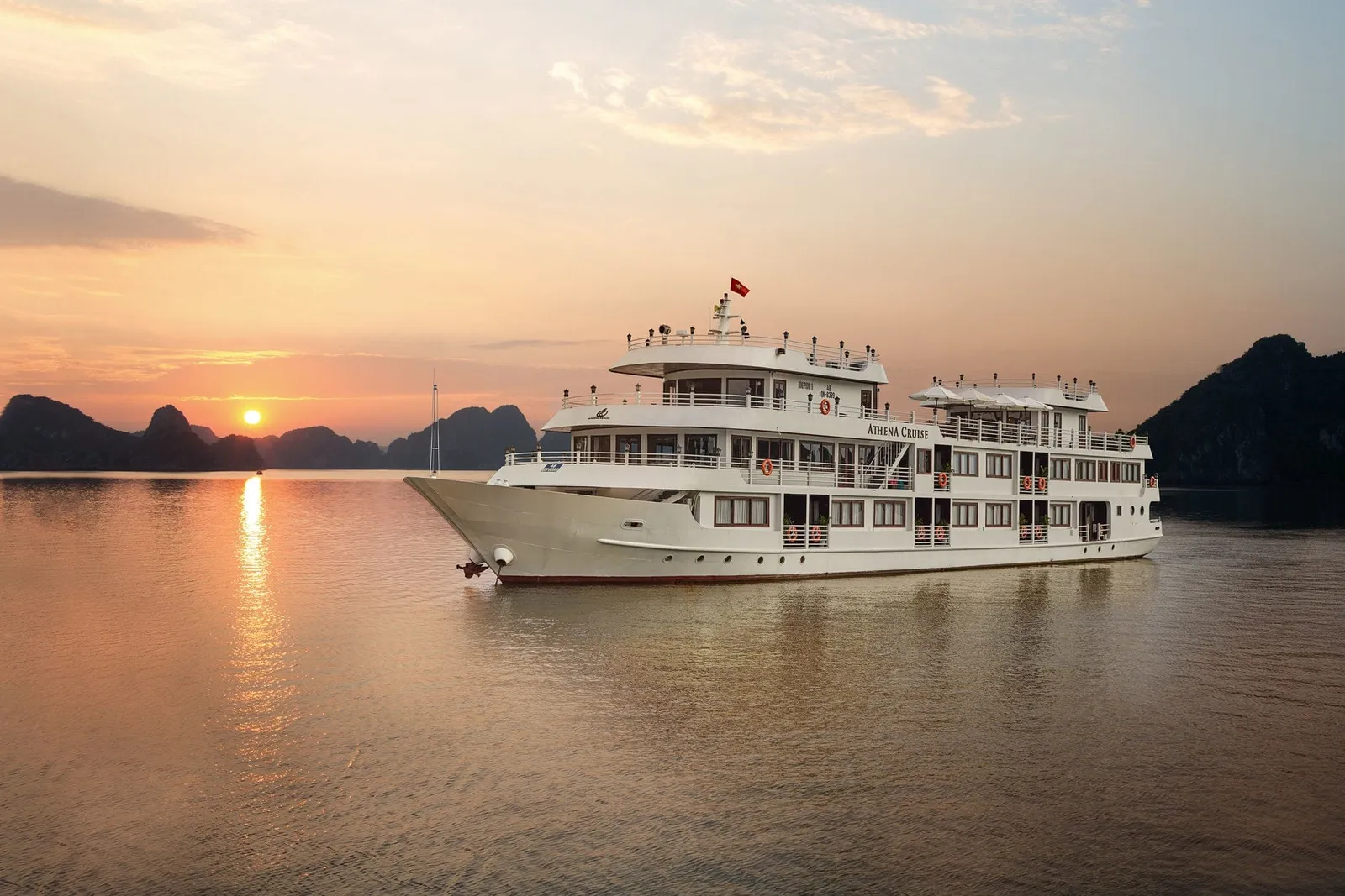 A white multi-deck steel cruise ship with a Vietnamese flag sits on calm water at sunset with limestone karst islands and a low sun in the background.