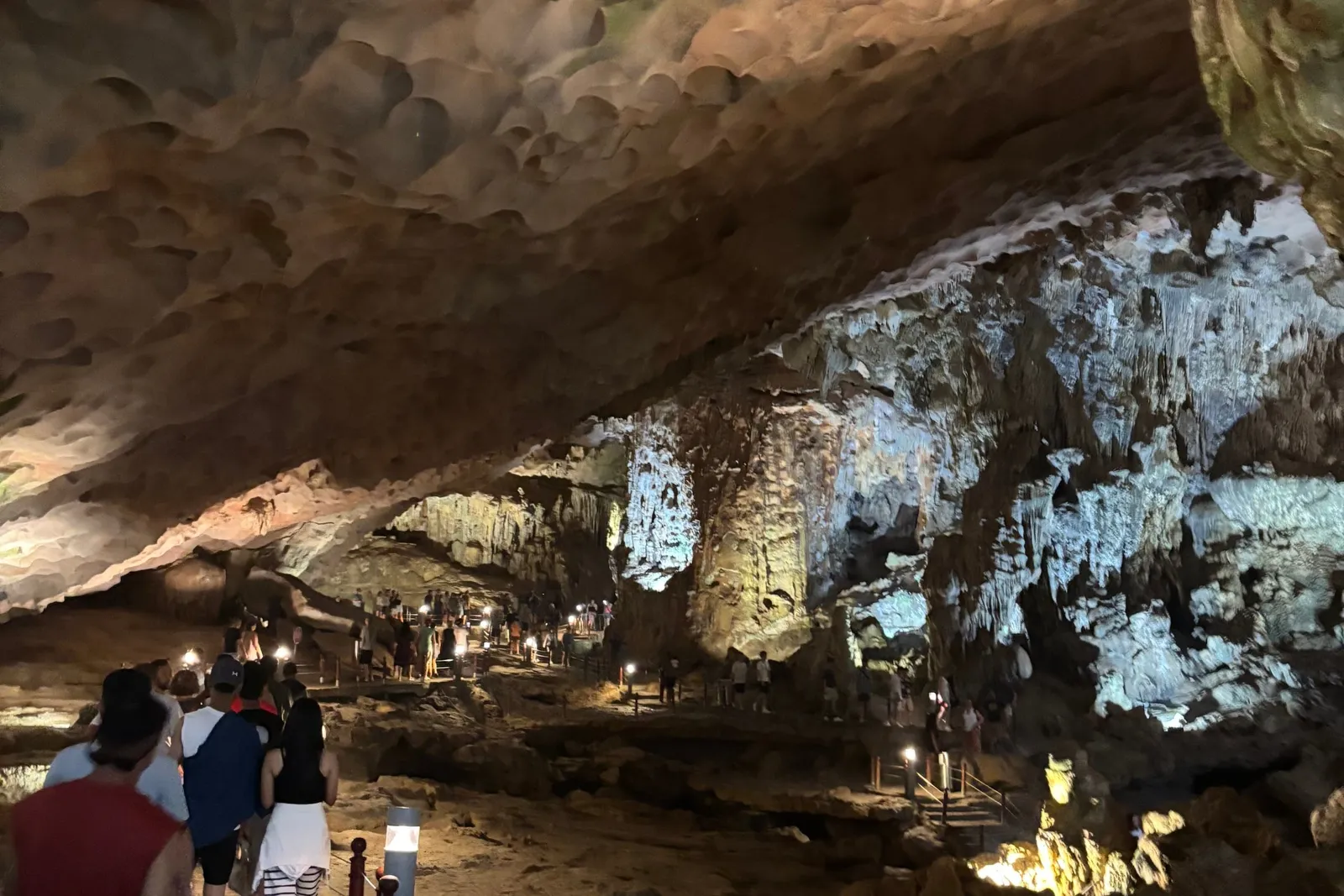 Tourists walk along a roped, lit path inside a large limestone cave with a honeycomb-textured ceiling and illuminated stalactite formations.