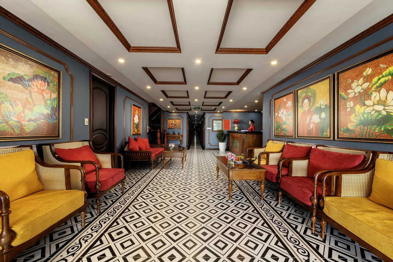 A long indoor lounge with black-and-white geometric tile floor, wooden rattan sofas with yellow and red cushions, lotus paintings on blue walls, and a receptionist in red at a wooden desk at the far end of a corridor.