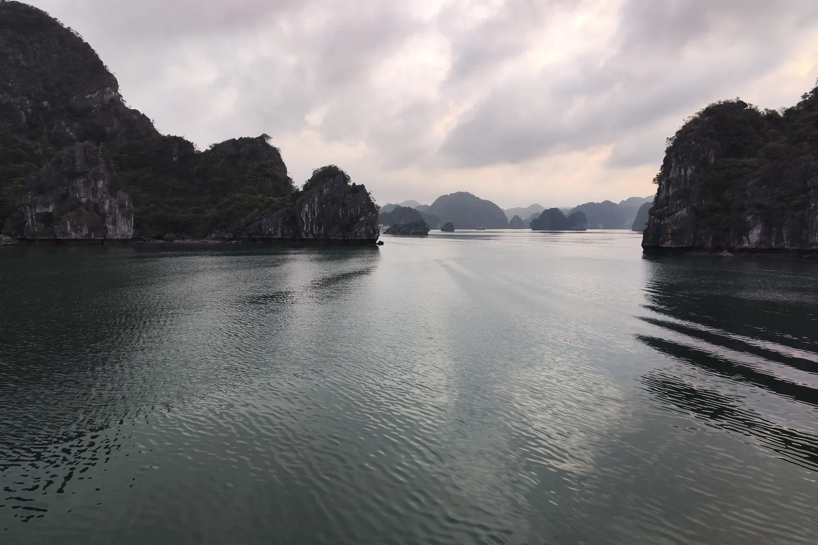 Calm green-grey water between forested limestone karst islands under an overcast pink-tinged sky, with ripples and a faint wake suggesting the photo was taken from a moving boat.