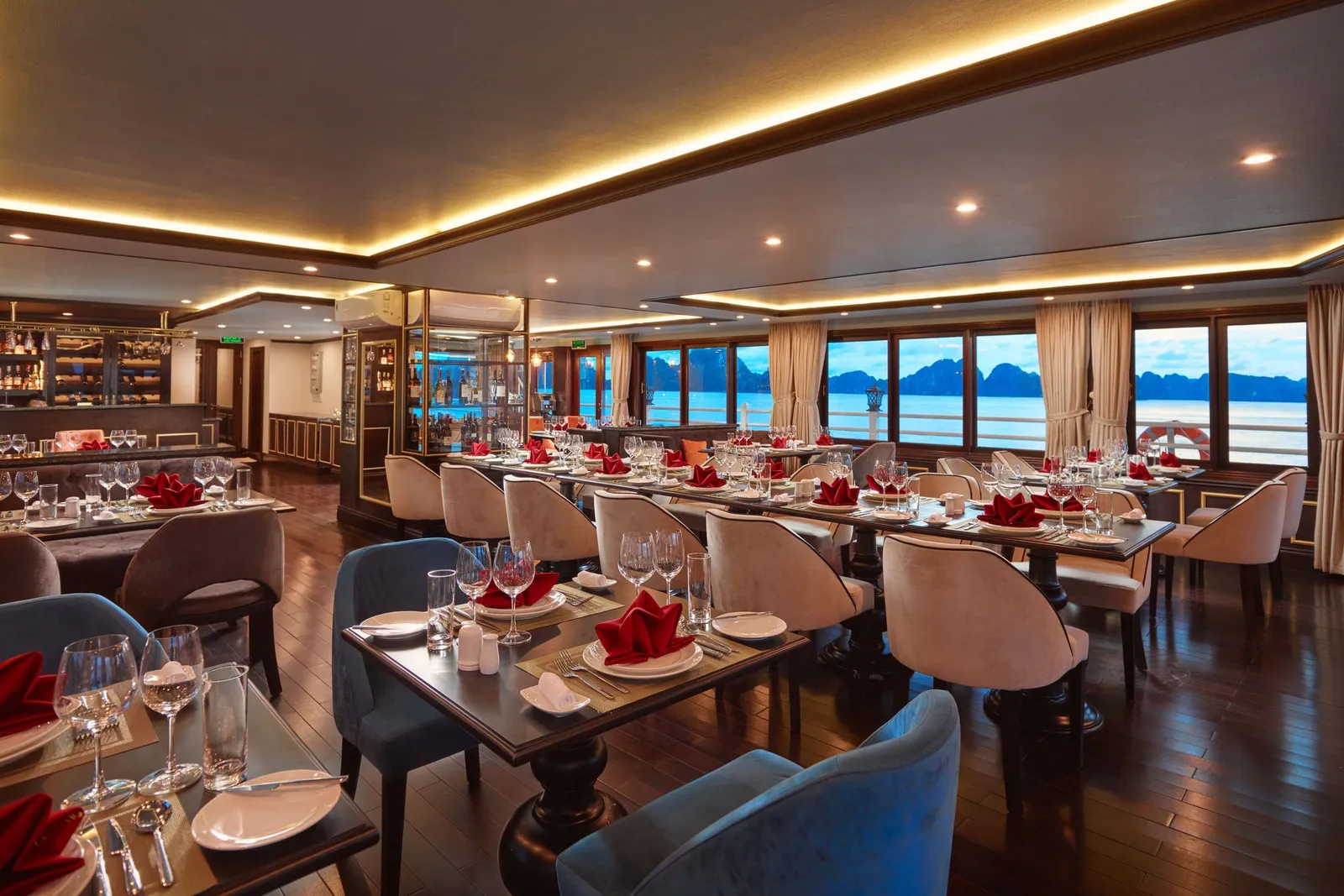 A boat's interior dining room with set tables, red folded napkins, wine glasses and upholstered chairs, with large windows showing calm water and limestone karst islands outside.