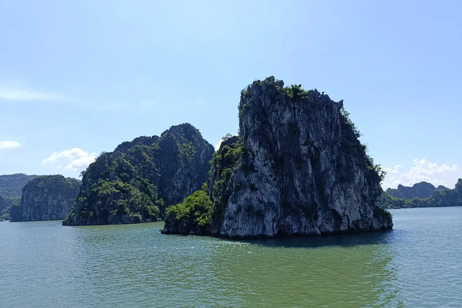 Limestone karst islands with green vegetation rise from calm green-blue water under a mostly clear blue sky, photographed from water level.