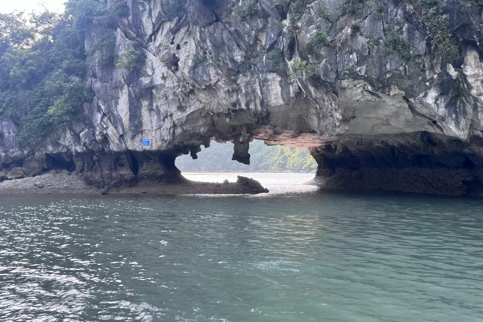 A limestone karst cliff with a natural arch opening onto a view of green hills beyond, above calm blue-green water, with a small blue sign on the rock and no overlaid text.