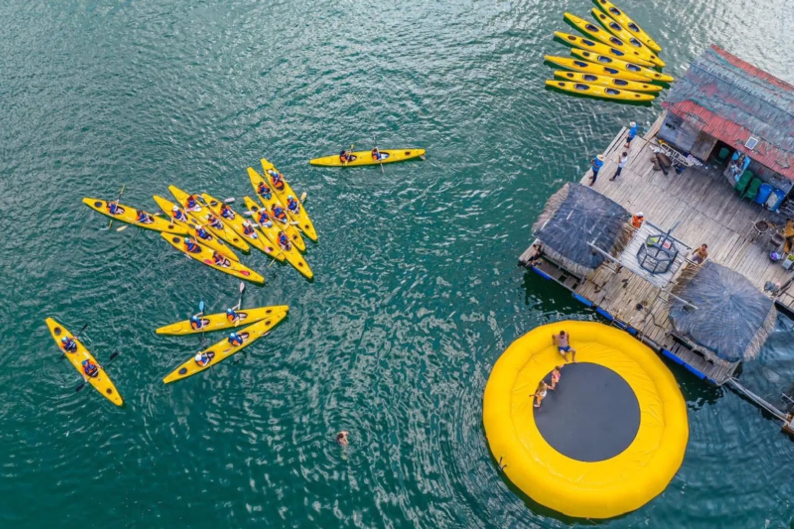 Aerial view of many yellow kayaks with paddlers on green water beside a floating wooden platform with thatched umbrellas and a round yellow inflatable trampoline with people on it.