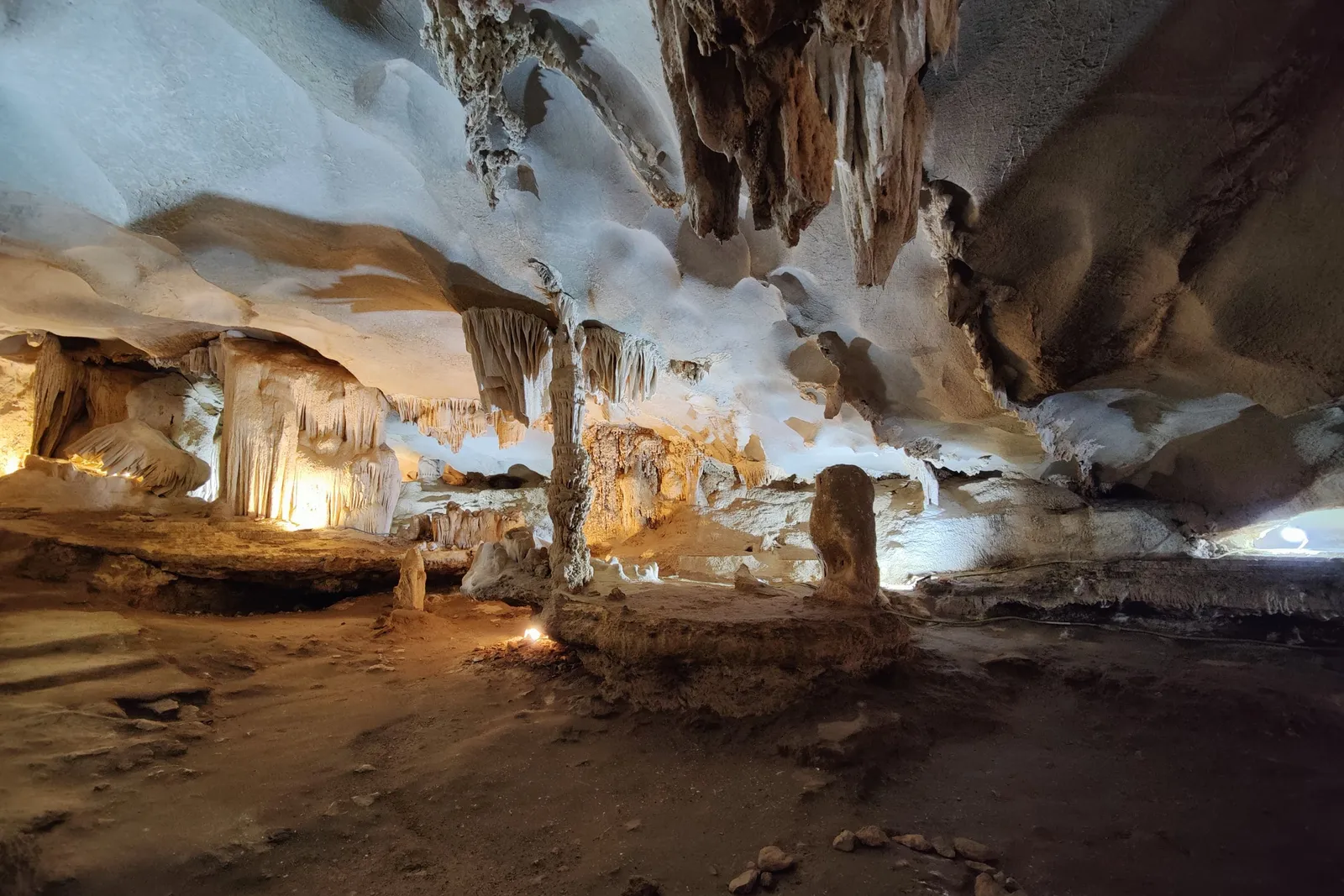 The interior of a limestone cave with stalactites, stalagmites and flowstone lit by warm lights, above a dirt floor scattered with small rocks.