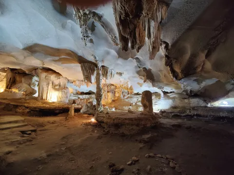 The interior of a limestone cave with stalactites, stalagmites and flowstone lit by warm lights, above a dirt floor scattered with small rocks.