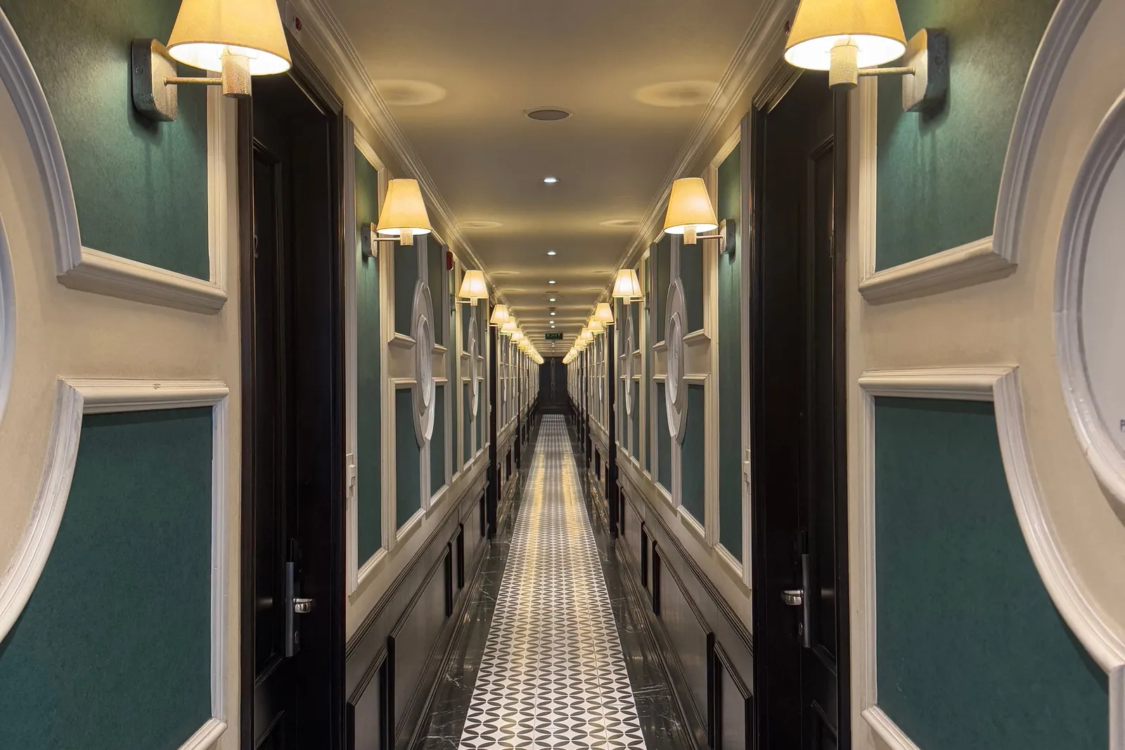 A very long, symmetrical indoor corridor with dark wooden doors, green wall panels with white moulding, lit wall sconces, and a black-and-white patterned tile runner on a dark marble floor.