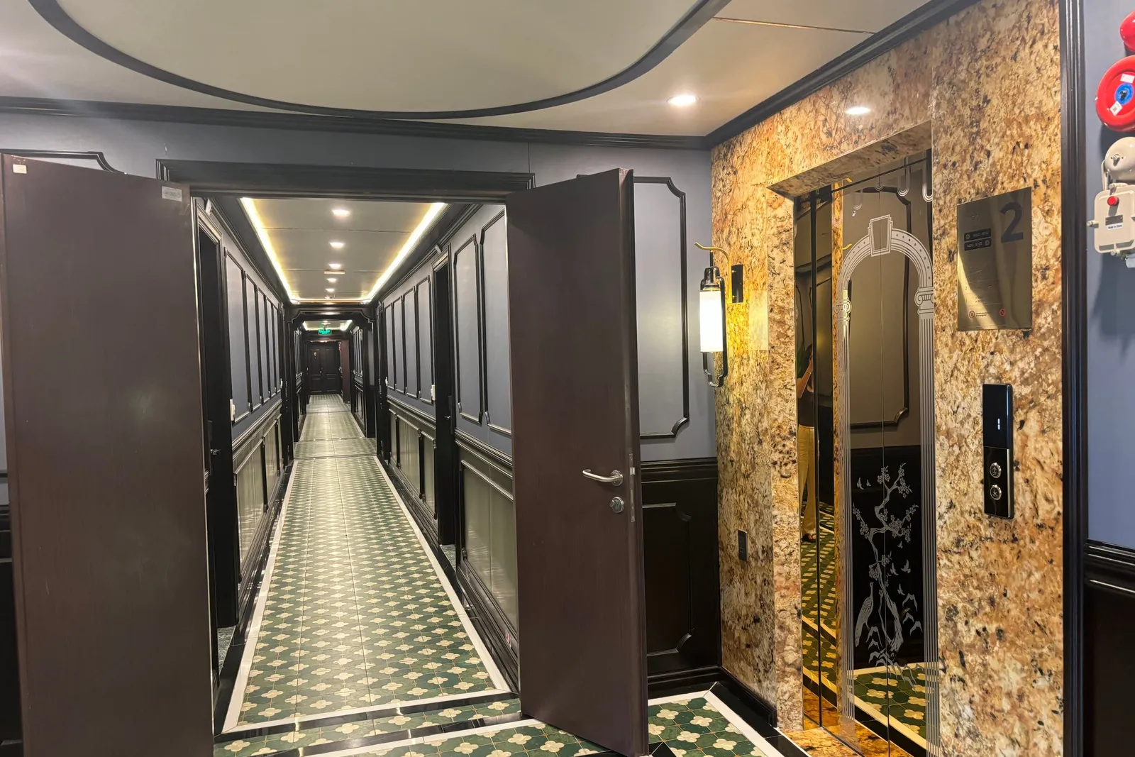 A long interior corridor with green patterned tile floor, dark wood doors and navy paneled walls, next to a granite-clad elevator marked with the number 2.