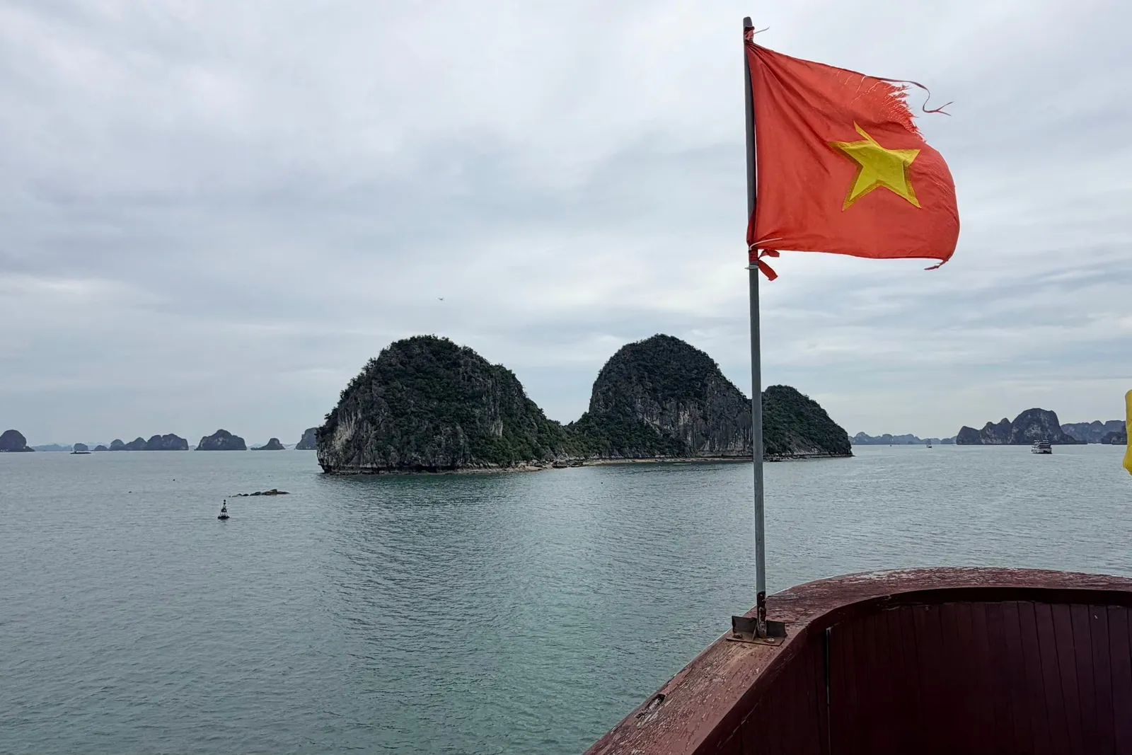 A Vietnamese flag on a pole at the bow of a boat with a dark red wooden rail, looking out over calm green water toward forested limestone islands under an overcast sky.