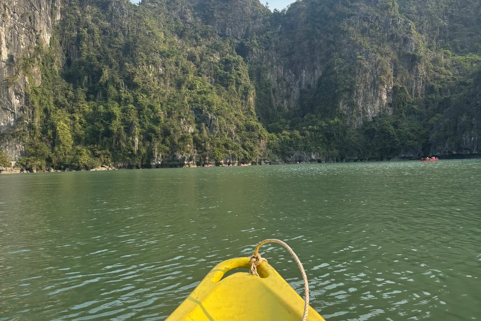 A first-person view from a yellow kayak with a person's sandal-clad feet in the foreground, looking across calm green water toward steep forested limestone karst cliffs under a clear sky.