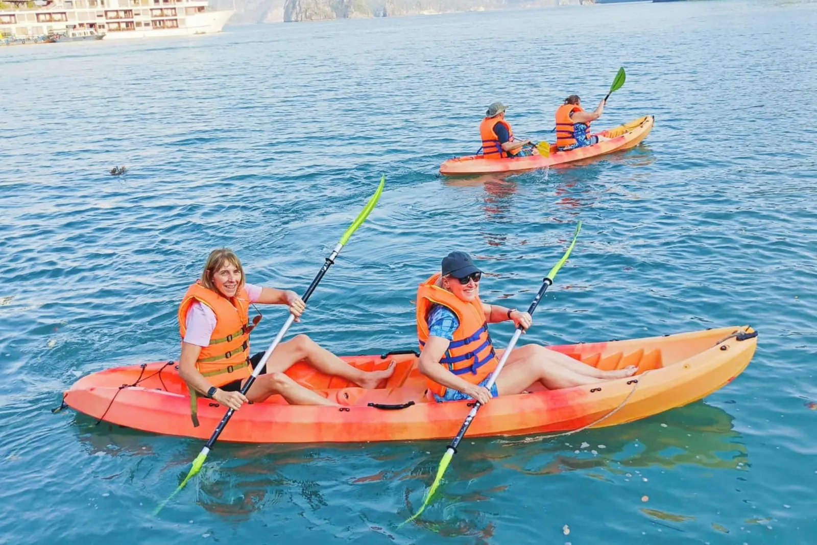 Two women in orange life jackets paddle an orange tandem kayak on blue-green water, with a second kayak with two paddlers behind them and a cruise ship and limestone cliffs in the distance.