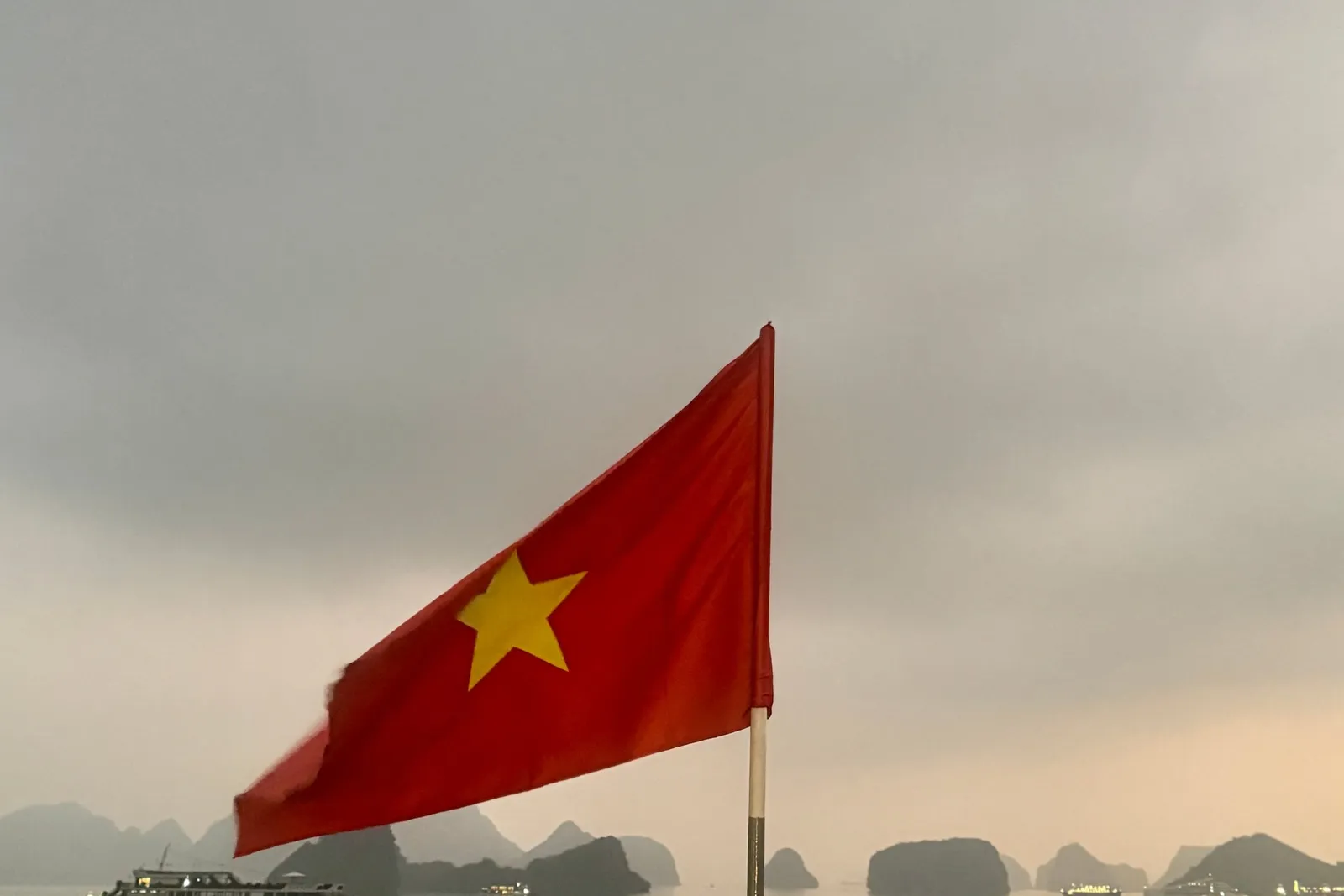 A Vietnamese flag on a pole in the foreground, seen from a boat deck railing, with limestone karst islands and a lit cruise boat on calm water under an overcast dusk sky.