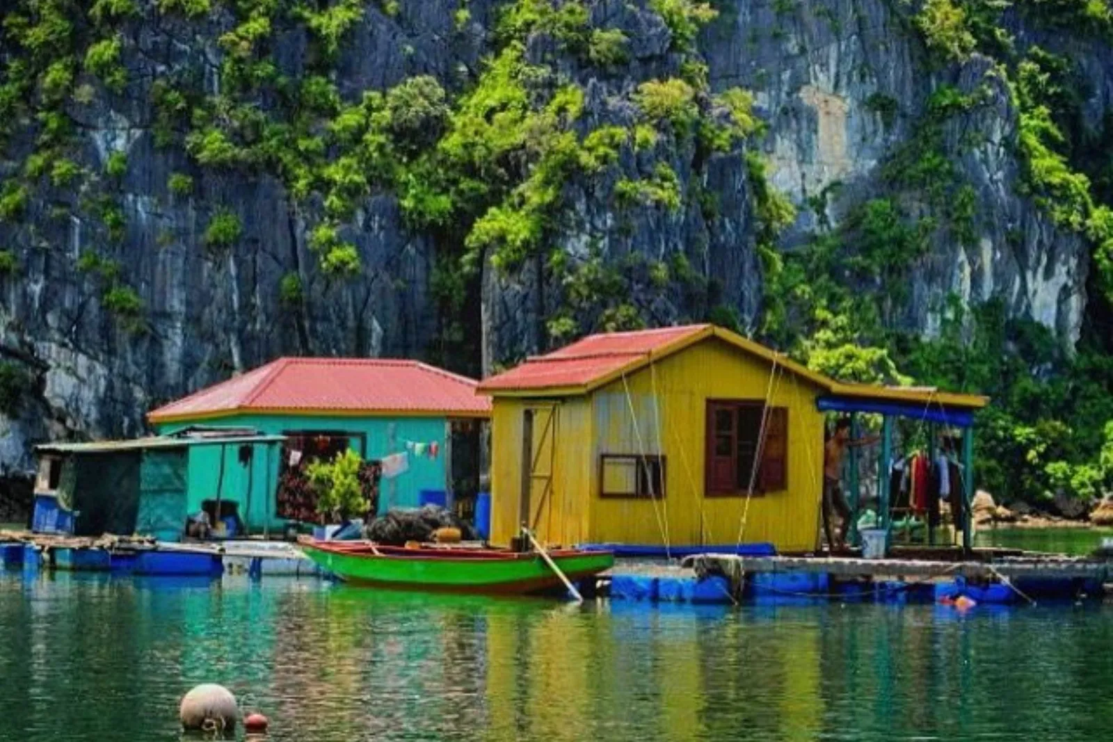 Colorful floating houses and a small green rowboat on calm green water in front of a steep limestone cliff covered in vegetation, with a person standing on the platform.