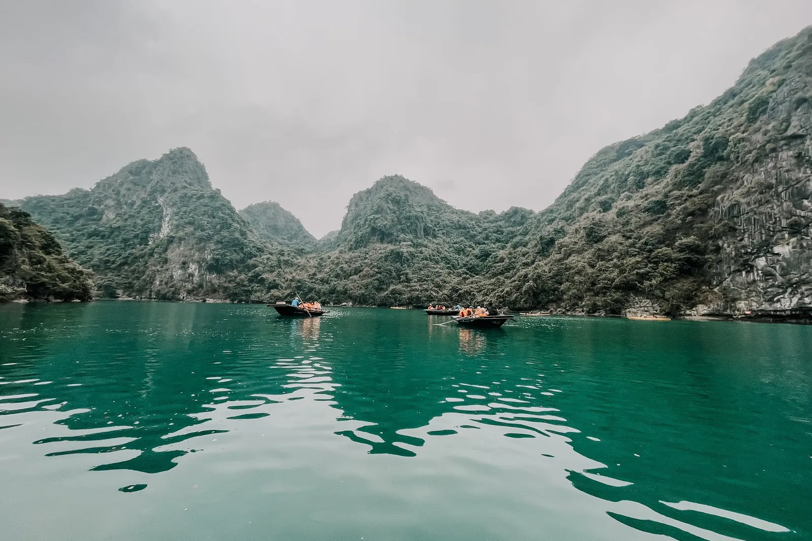 Green-turquoise water in a bay surrounded by forested limestone karst hills under an overcast sky, with small rowing boats carrying people in orange life jackets in the distance.