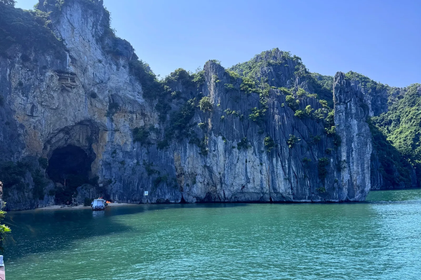 Turquoise water in front of a steep limestone karst cliff with a dark cave opening and a small white boat near a sandy beach, shot from a boat whose edge is visible at the lower left under a clear blue sky.
