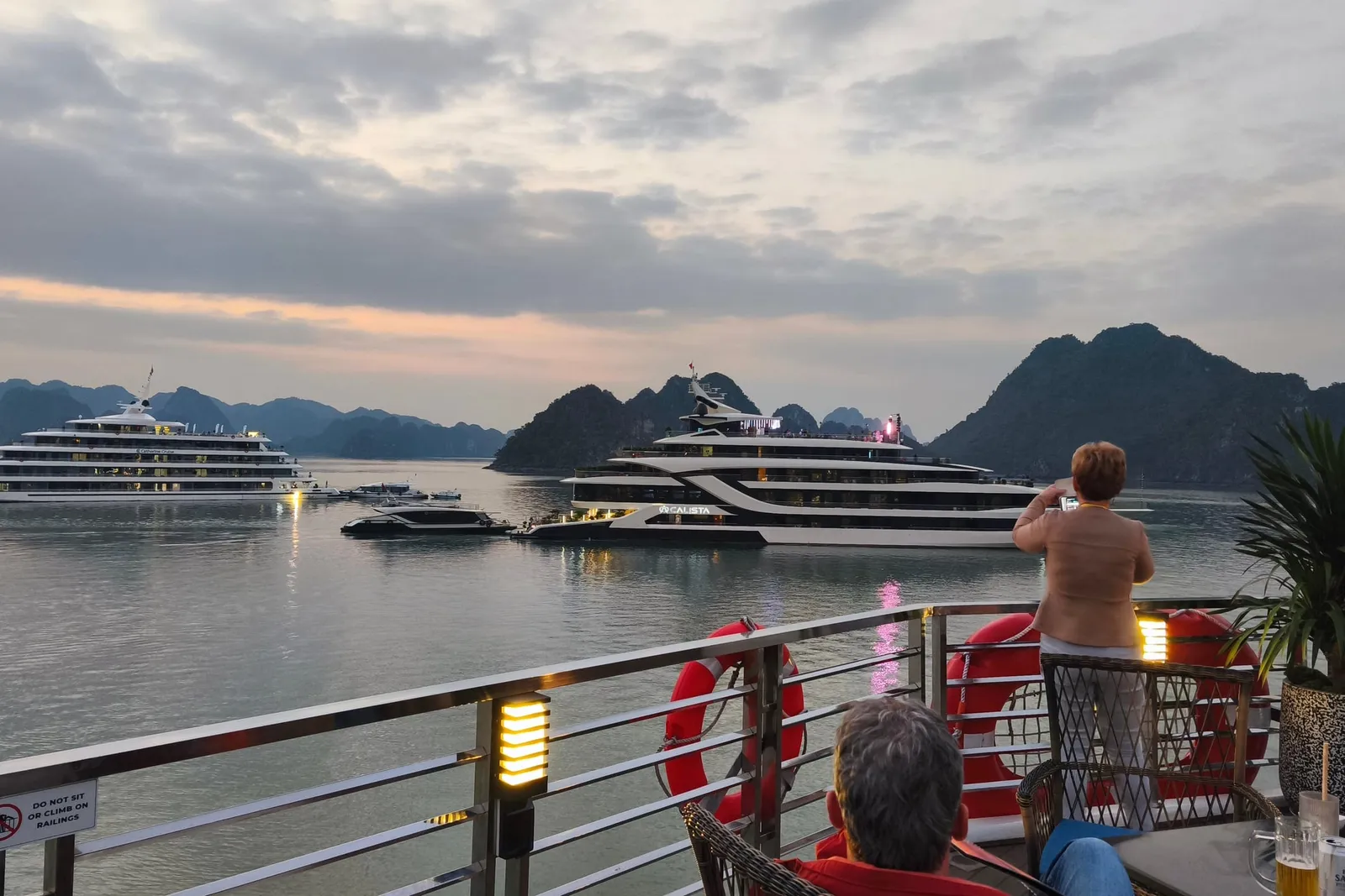 From a railed upper deck at dusk with red lifebuoys, a potted plant and a table with drinks, a woman stands photographing two large white cruise ships on calm water among limestone karst islands, while a seated person is seen from behind in the foreground.