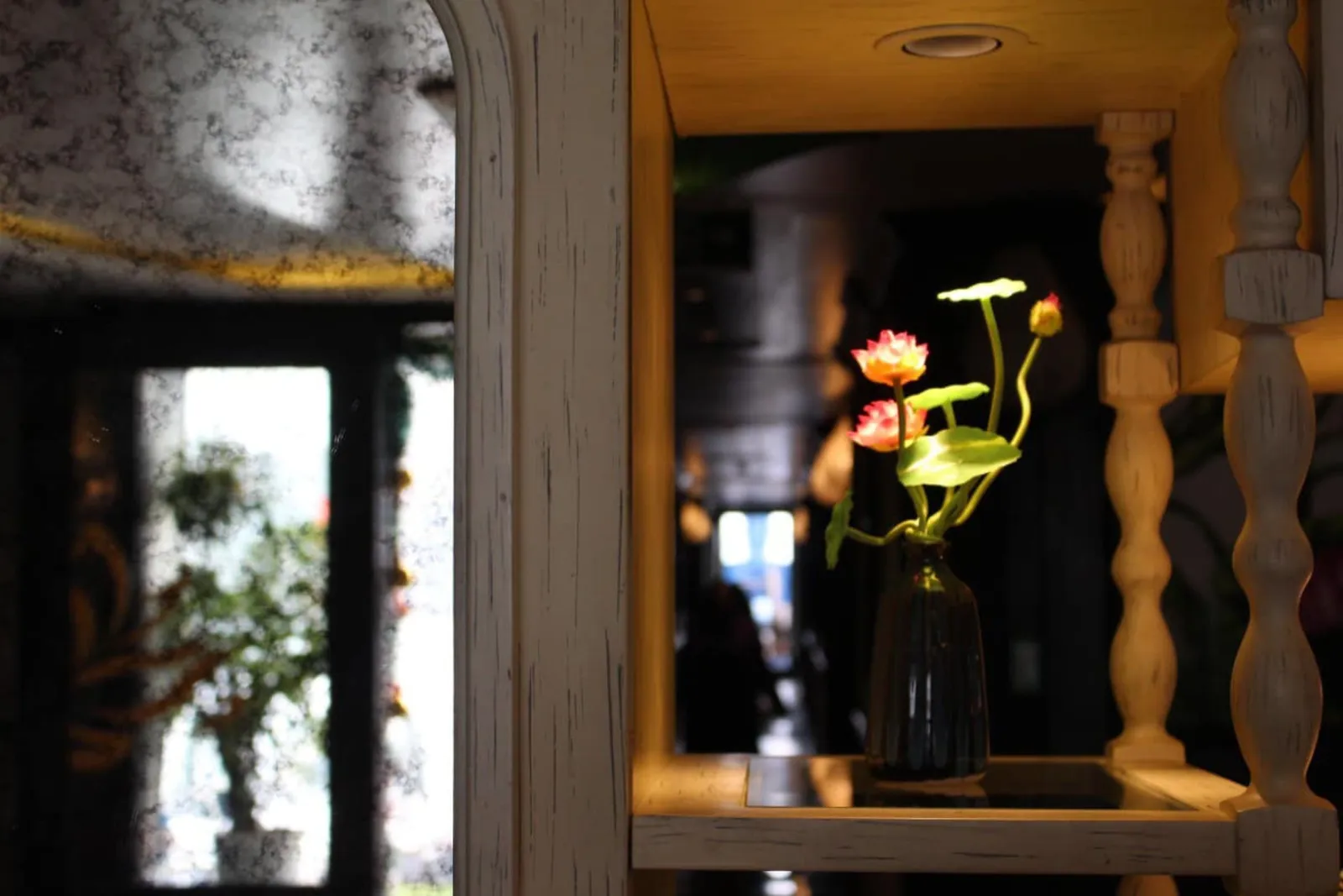 A dark glass vase holding pink lotus flowers and buds sits on a cream wooden shelf with turned spindles, beside an antiqued mirror reflecting a potted plant and a dim corridor.
