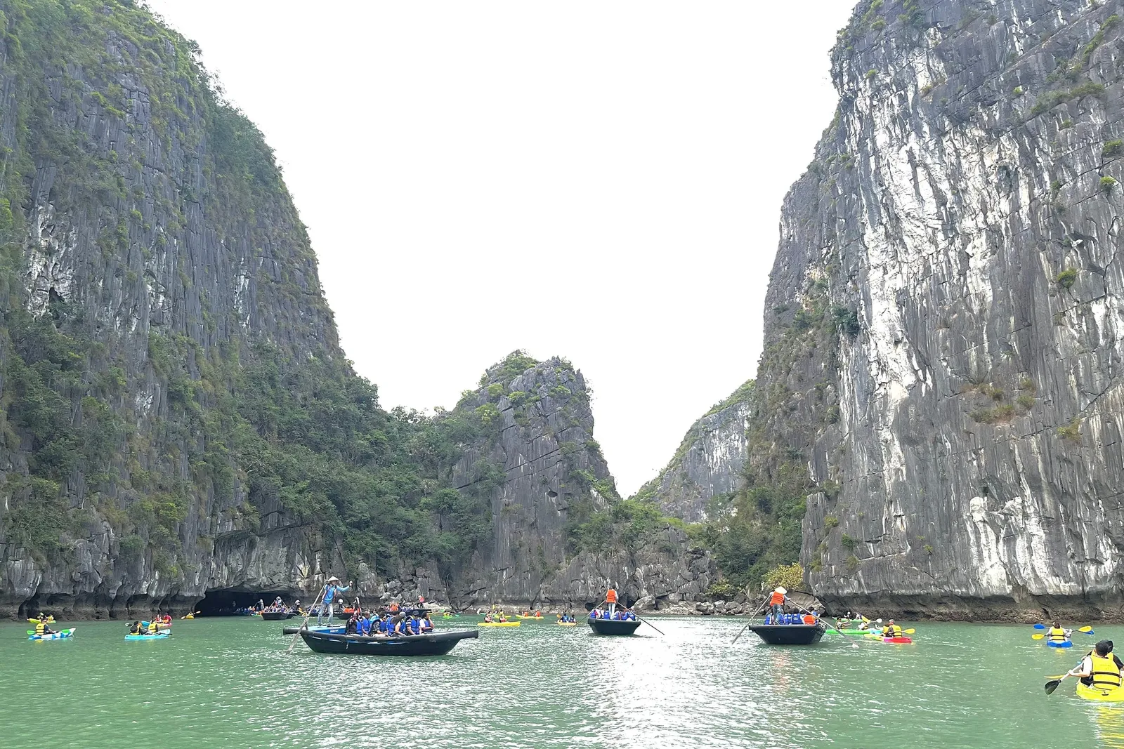 Green water between tall limestone cliffs with small rowing boats carrying passengers in blue vests and several kayakers, with a cave opening at the base of the left cliff.