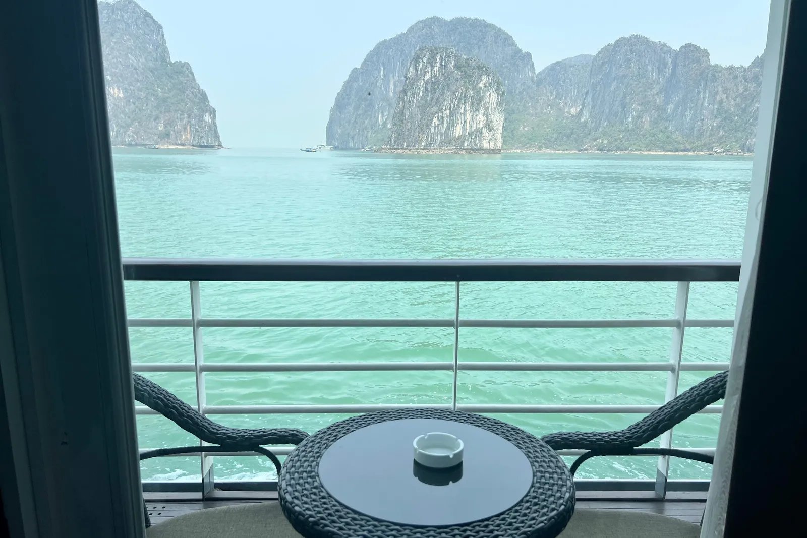 A view through an open balcony door onto a railing, a small wicker table with an ashtray and two chairs, with turquoise water and limestone karst islands beyond in a hazy sky.