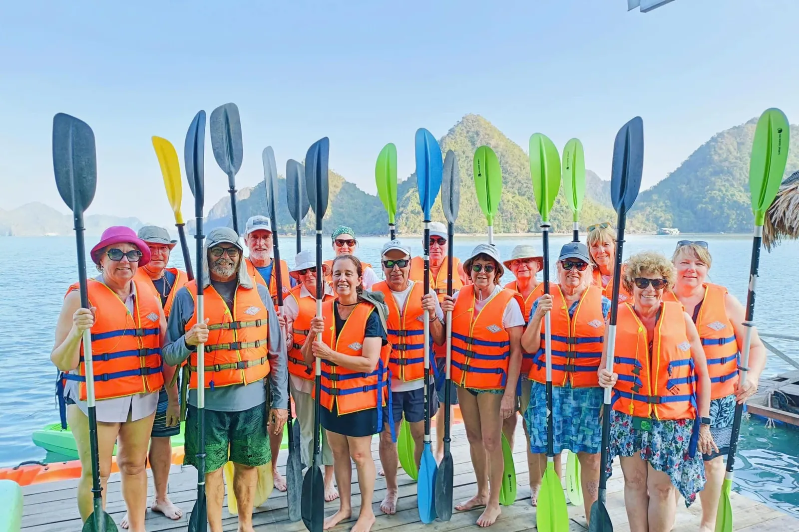 A group of about a dozen people in orange life jackets stand on a wooden floating platform holding upright kayak paddles, with calm water and green limestone karst hills behind them.