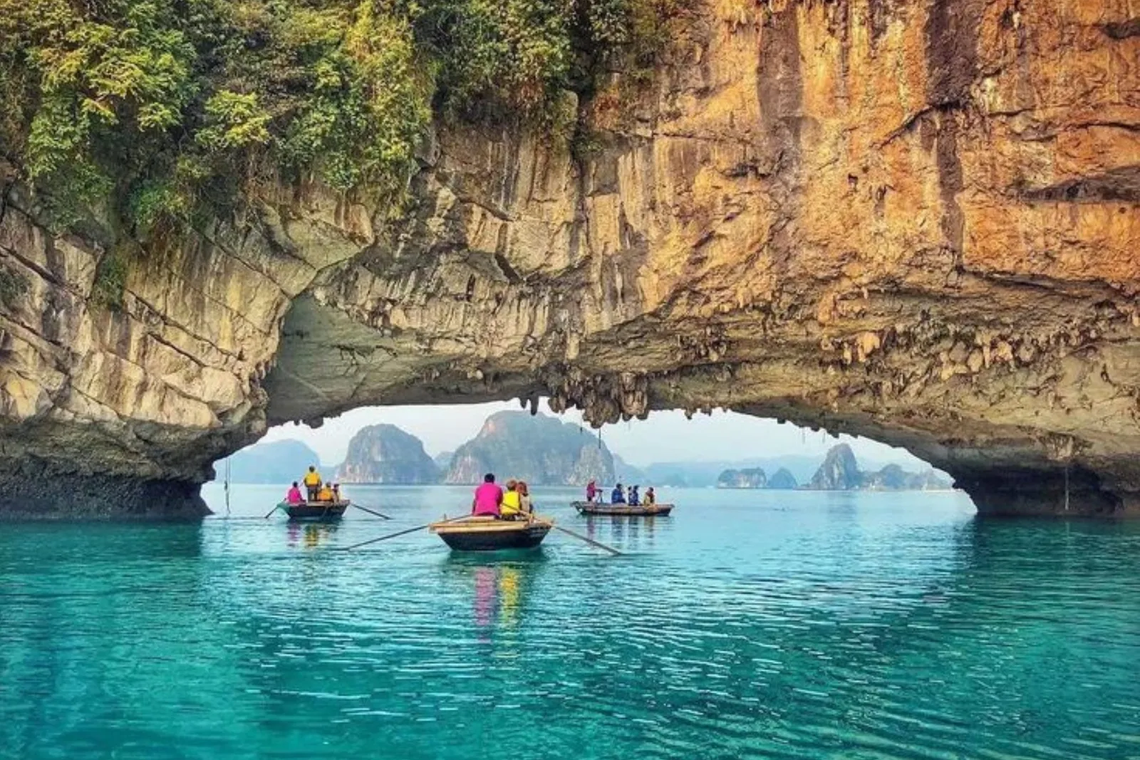Three small rowing boats with people in life vests glide on turquoise water toward a large limestone rock arch, with karst islands visible in the hazy distance beyond.