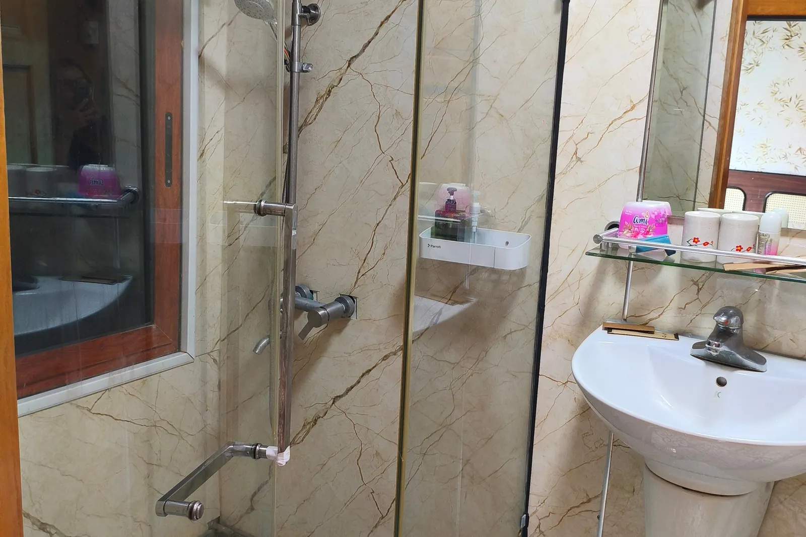 A small ensuite bathroom with a glass-enclosed shower, marble-look wall panels, patterned floor tiles, a wall-mounted sink with a mirror and shelf of toiletries, and a wood-framed interior window.