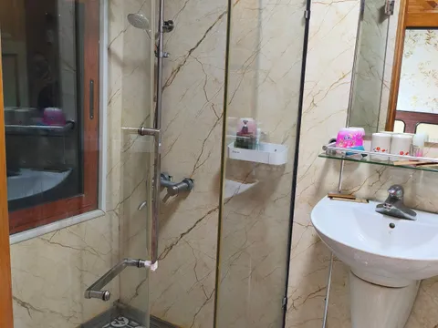 A small ensuite bathroom with a glass-enclosed shower, marble-look wall panels, patterned floor tiles, a wall-mounted sink with a mirror and shelf of toiletries, and a wood-framed interior window.