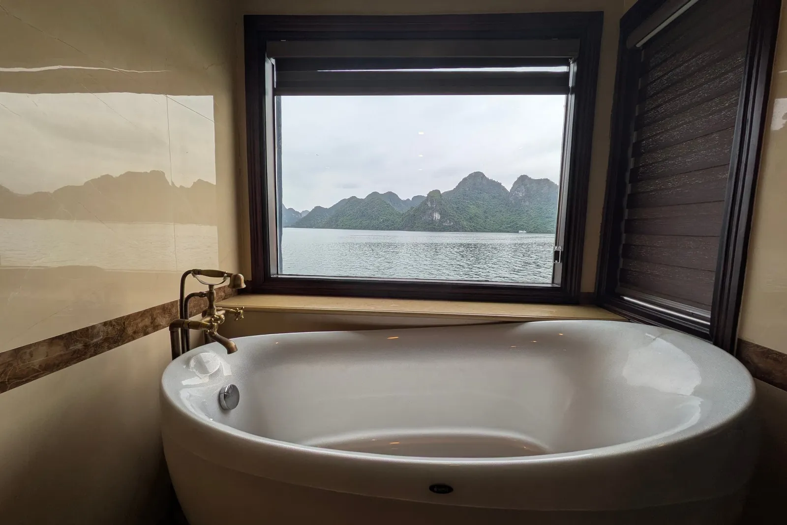 A white freestanding oval bathtub with a brass tap sits under a large window that looks out over calm water toward green limestone karst islands under an overcast sky.