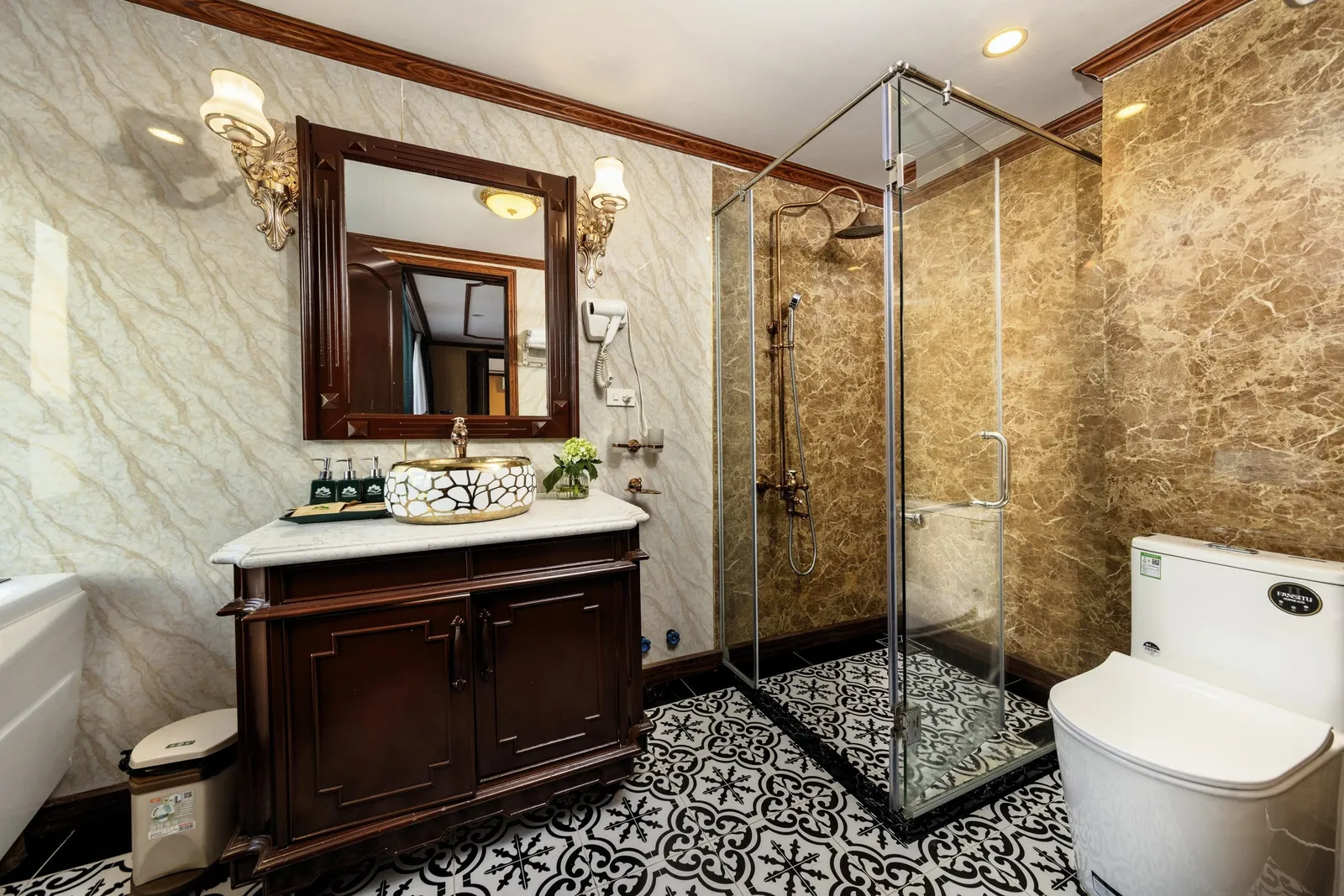 A marble-walled ensuite bathroom with a dark wood vanity, gold patterned vessel sink, framed mirror with wall lamps, glass shower enclosure, patterned black-and-white tile floor, and a white toilet with a small manufacturer logo on the tank.