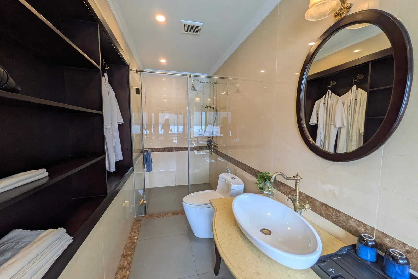 A bathroom with a glass-enclosed shower, toilet, oval vessel sink with antique-style brass tap, round dark-framed mirror, and dark open shelving holding folded towels and hanging white bathrobes.