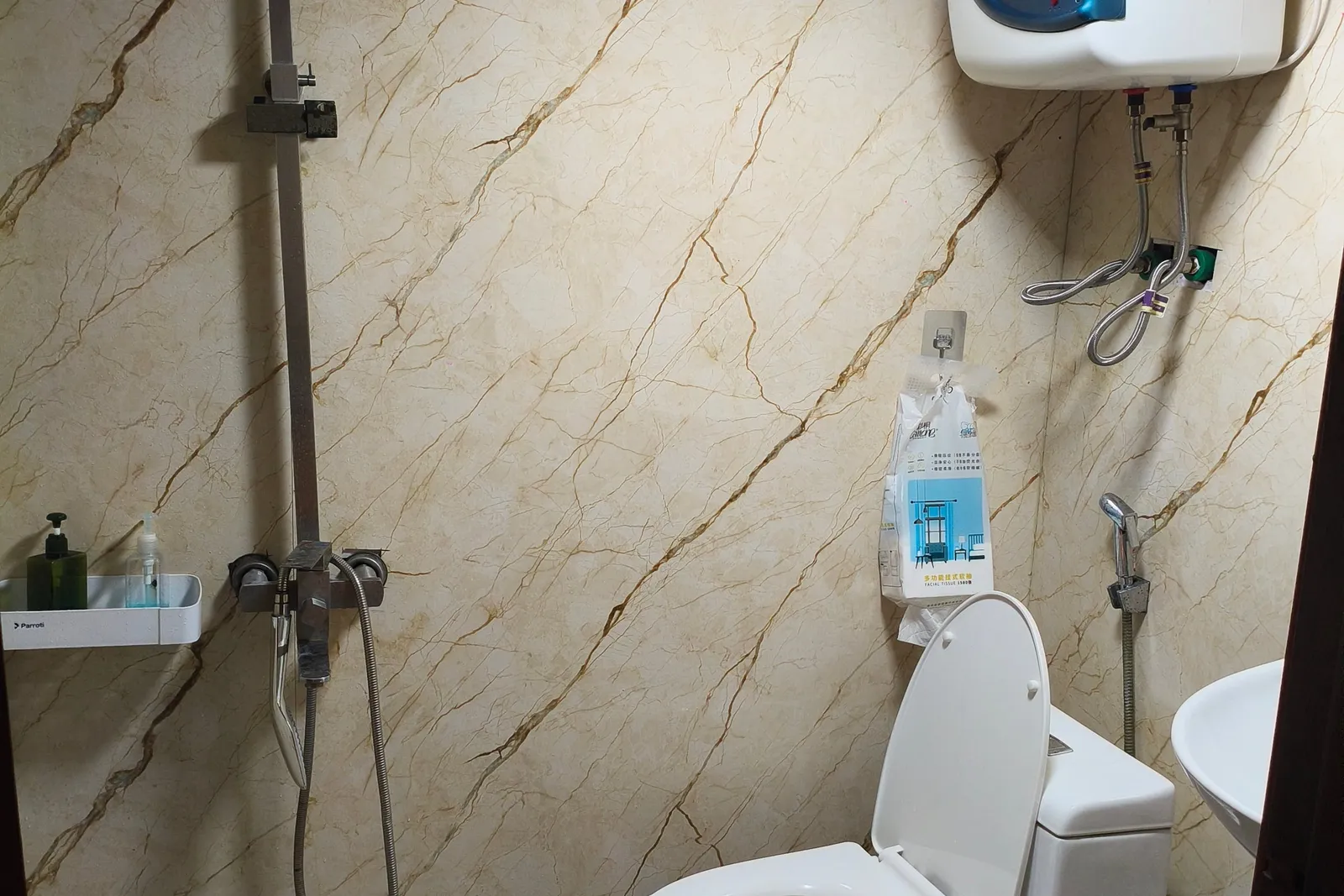 A small bathroom with marble-look wall panels, patterned floor tiles, a white toilet, a wall-mounted Olympic water heater, a shower fixture and a shelf with toiletries, with product brand names printed on the equipment.