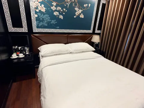 A double bed with white linens in a cabin with a dark panelled wall, a blue magnolia-and-bird mural with Greek-key borders, brown curtains, a tea set on a side table, and a wooden floor.