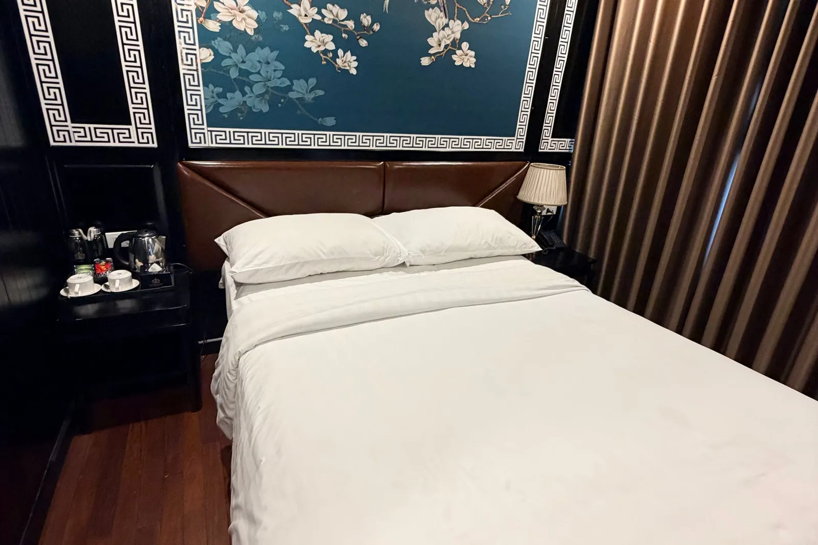A double bed with white linens in a cabin with a dark panelled wall, a blue magnolia-and-bird mural with Greek-key borders, brown curtains, a tea set on a side table, and a wooden floor.