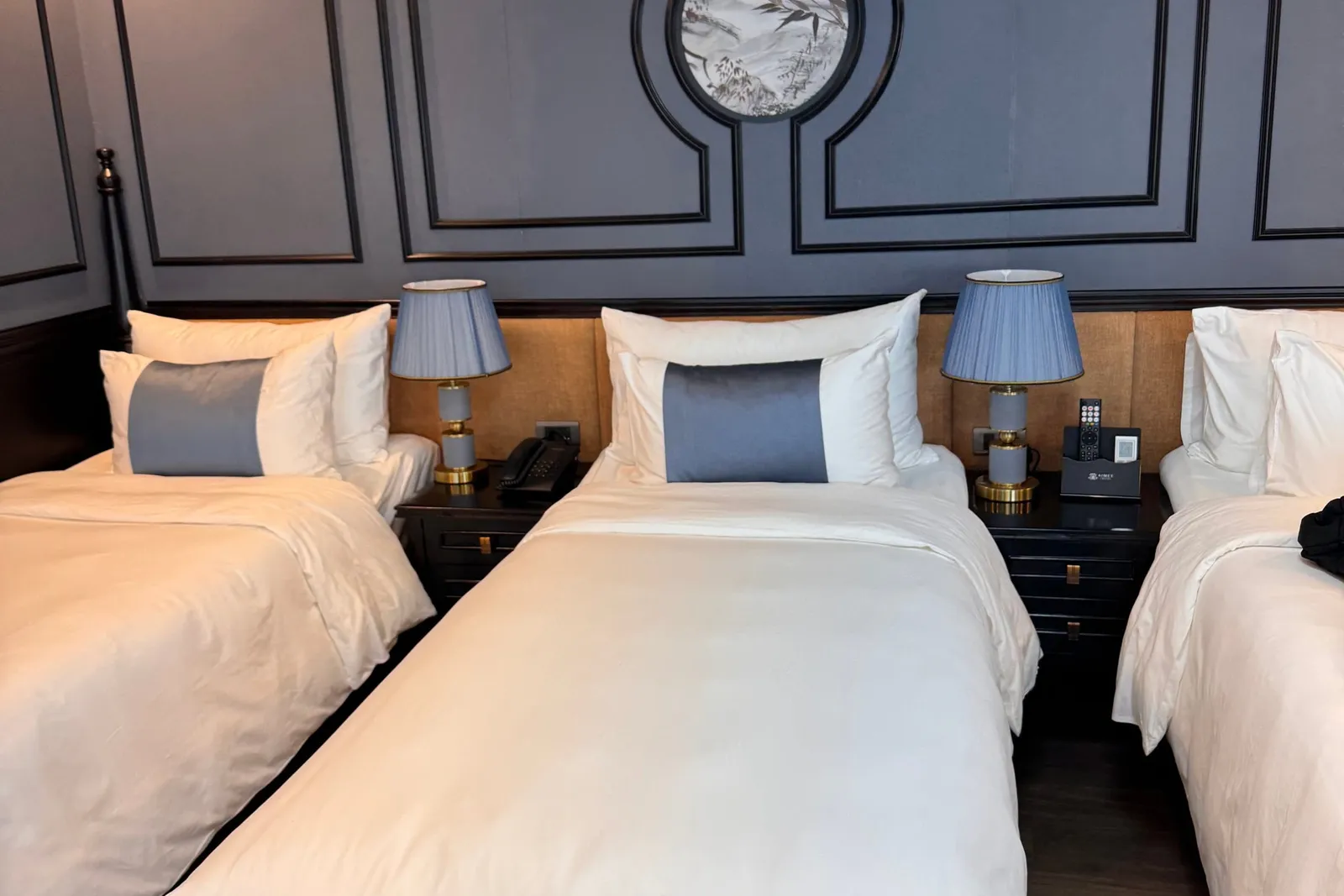 Three twin beds with white linens and blue-grey bolster cushions sit against a dark grey paneled wall with a round bird-and-landscape painting, flanked by bedside tables with blue-shaded lamps, a phone, and a small branded remote holder.