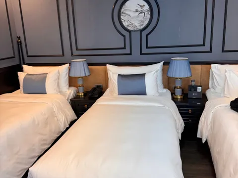 Three twin beds with white linens and blue-grey bolster cushions sit against a dark grey paneled wall with a round bird-and-landscape painting, flanked by bedside tables with blue-shaded lamps, a phone, and a small branded remote holder.