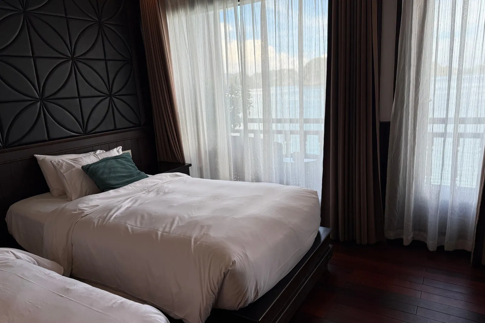 A dim cabin with two twin beds in white linens, a teal cushion, a dark geometric padded headboard panel, sheer and brown curtains, and a balcony railing with water and distant karst-like hills visible outside.