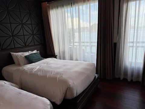 A dim cabin with two twin beds in white linens, a teal cushion, a dark geometric padded headboard panel, sheer and brown curtains, and a balcony railing with water and distant karst-like hills visible outside.
